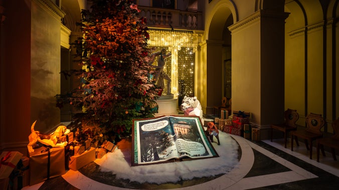a photo of shadow the snow leopard behind a large story book. to the left is a christmas tree and behind is a fairy lights curtain